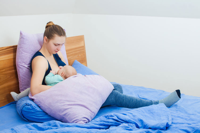 mother sitting in bed surrounded by cushions breastfeeding her baby