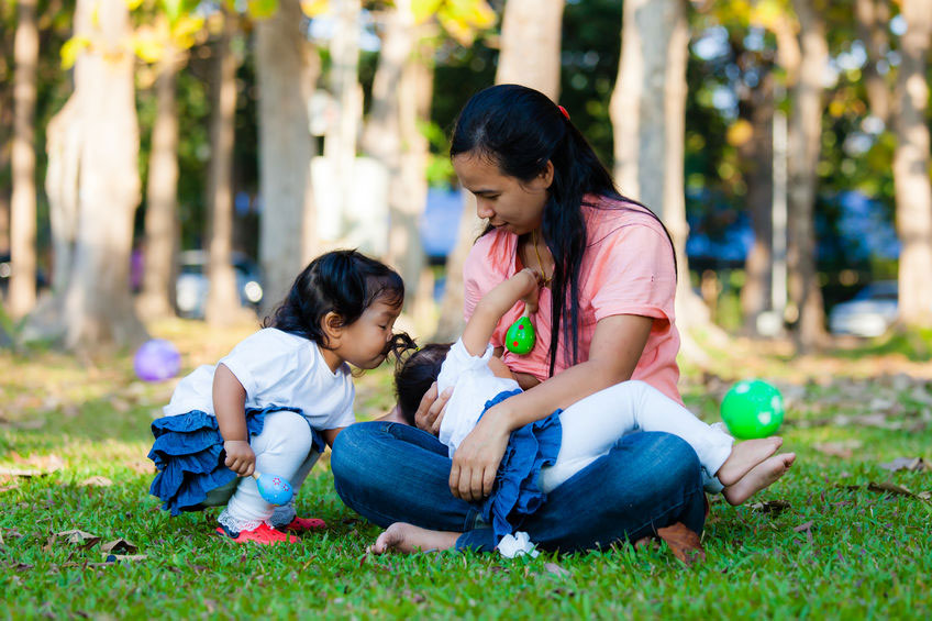 Mother sitting cross legged in park breastfeeding a toddler, her sister wants to play